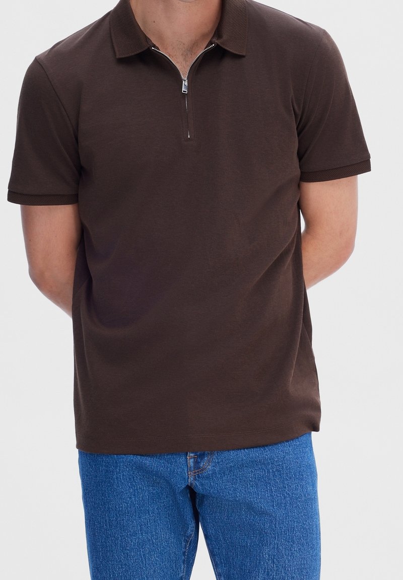 Homme portant un polo marron à manches courtes avec une fermeture éclair à l'avant et un jean bleu, debout les mains dans le dos.