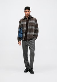 Νεαρός άνδρας φοράει καφέ πλακωτό πουκάμισο με μπουφάν, σκούρο καφέ πουκάμισο, γκρίζες παντελόνες, μαύρα παπούτσια, κρατώντας ένα διπλωμένο μπλε μπουφάν.