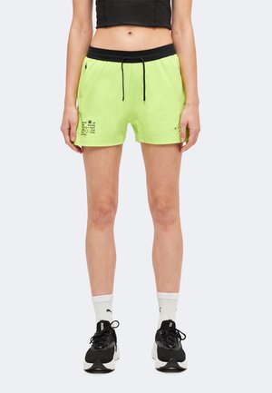 Personne portant un short vert fluo à cordon, un crop top noir, des chaussettes blanches Puma et des baskets noires Puma, debout les bras détendus le long du corps.