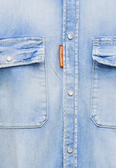 Μπροστινό μέρος ανοιχτό μπλε πουκάμισου από denim με δύο τσέπες με καπάκι, επάργυρα κουμπιά κλικ και πορτοκαλί υφασμάτινη ετικέτα με την ένδειξη "DSQUARED2" ανάμεσα στις τσέπες.