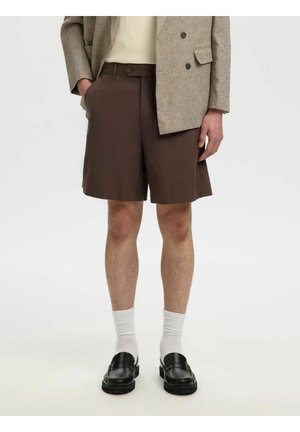 Person trägt einen beigen Blazer, ein cremefarbenes Oberteil, braune maßgeschneiderte Shorts, weiße Socken und schwarze Loafer und steht vor einem schlichten Hintergrund.