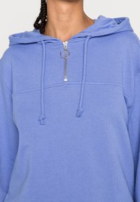 Ljusblå hoodie med dragkedjekrage med ett silverring, snörningsdetaljer och en strukturerad tyg. Avslappnad passform och avslappnad design.