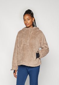 Beiger Fleece-Pullover mit Kapuze, ausgestattet mit einer Fronttasche und schwarzen Akzenten an den Ärmeln. Weicher Stoff, lockere Passform und abgerundeter Saum.