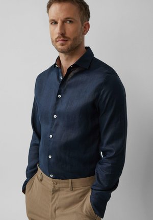 Navy blue overhemd met lange mouwen van lichte stof, voorzien van witte knopen en een gemusterde kraag. Gecombineerd met beige broeken, die een verzorgde pasvorm laten zien.