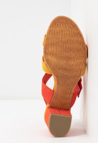 Sandale femme avec brides en daim orange et jaune et petit talon bloc, vue par la semelle sur fond blanc.