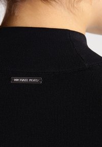 Schwarzer gerippter Pulloverkragen mit einem silbernen rechteckigen Michael Kors-Logoschild auf der Rückseite. Der Stoff hat eine strukturierte Optik.
