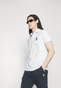 Hvit polo-skjorte med korte ermer, med en svart grafisk design på brystet. Kombinert med marineblå bukser og svarte solbriller.