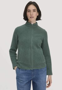 Groene fleecejack met een hoge kraag en ritssluiting aan de voorkant, voorzien van twee zijzakken en een subtiele textuur. Gepaard met blauwe denim jeans.