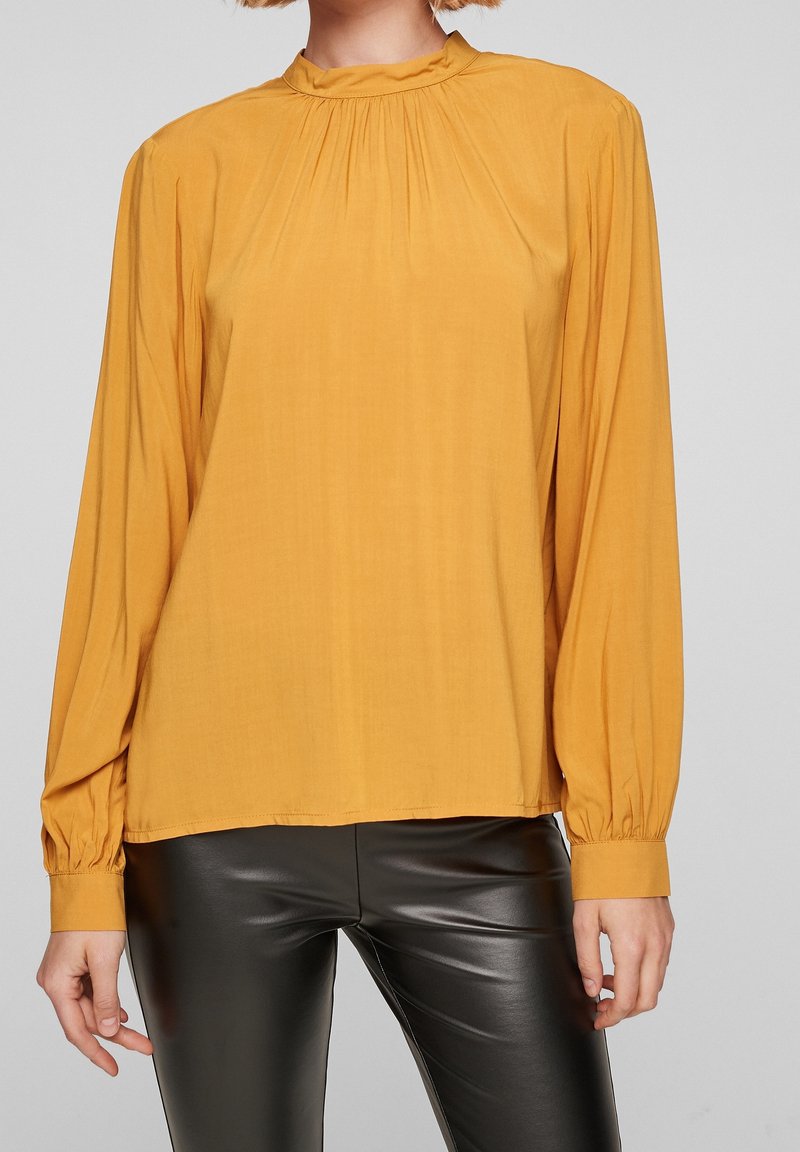 Blusa giallo senape con colletto alto, maniche lunghe a palloncino e dettagli arricciati al collo. Tessuto morbido e vestibilità comoda.