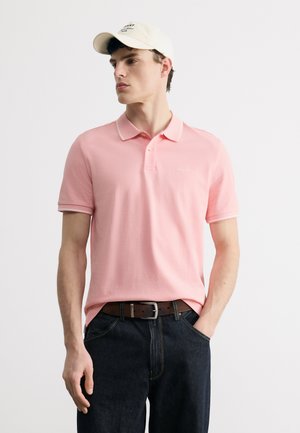 Giovane uomo con una polo rosa chiaro, jeans scuri, cintura marrone e un berretto bianco, in piedi con una mano in tasca e lo sguardo rivolto a sinistra.