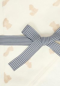 Koc bawełniany w kremowej podstawie z wzorem w jasnobrązowe misie pluszowe, wykończony granatową i białą wstążką zawiązaną w kokardę.