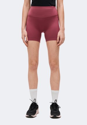 Personne portant un short de cycliste bordeaux taille haute, des chaussettes blanches montantes et des chaussures de sport noires, debout devant un fond uni.