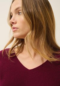 Burgundi värviline ribakudumine sviiter V-kaelusega, millel on tekstuuriline kangas ja lõtv sobivus. Juuksed langevad pehmete lainetena näo ümber.