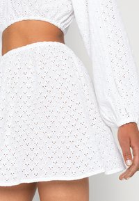 Vestido curto branco em tecido de ilhós com padrões florais, cintura franzida e mangas bufantes. Textura suave, design casual.