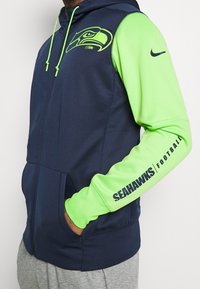 Navy zip-up hoodie with green sleeves, featuring a Seahawks logo on the chest and "SEAHAWKS FOOTBALL" printed on the arm. Smooth fabric, casual fit.
