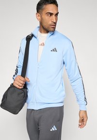 Casaco azul-claro com fecho éclair, detalhes em cinza, logótipo preto no peito, combinado com calças cinza, segurando uma pequena bolsa preta. Textura suave em toda a peça.