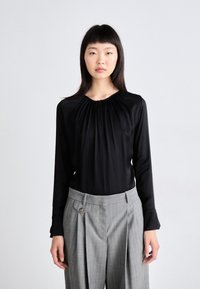 Mustat pitkähihaiset paita, jossa on rypytetty kaula-aukko, yhdistettynä harmaisiin raidallisiin korkeavyötäröisiin housuihin, joissa on pleatit ja nappiyksityiskohtia.