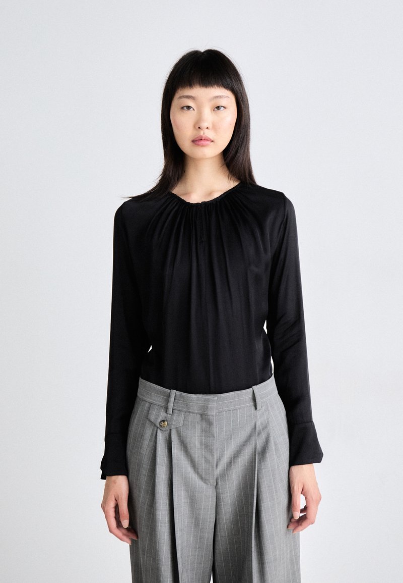 Mustat pitkähihaiset paita, jossa on rypytetty kaula-aukko, yhdistettynä harmaisiin raidallisiin korkeavyötäröisiin housuihin, joissa on pleatit ja nappiyksityiskohtia.