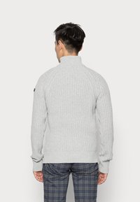 Pull gris clair côtelé avec un col montant et des manches longues. Comprend des épaules raglan et des poignets ajustés. Porté avec un pantalon à carreaux.