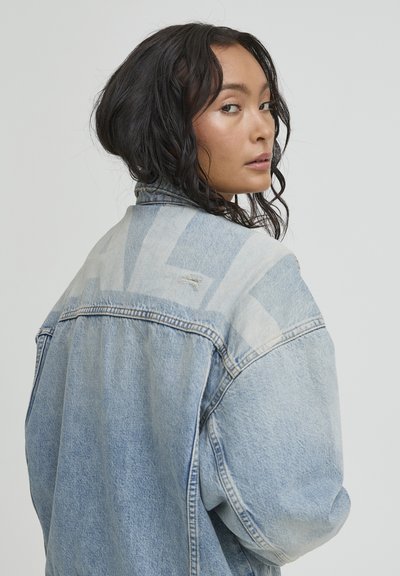 Woman with long dark curly hair looks over her shoulder wearing a light blue denim jacket against a plain background.