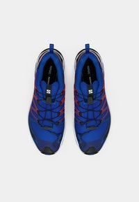Kék sportcipők dombornyomott szintetikus felsőrésszel, fekete részletekkel és fűzőrendszerrel, letisztult kialakítással és lekerekített orral.