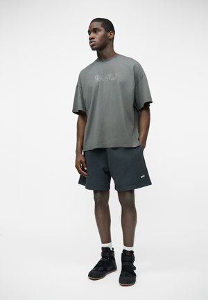 Mladi moški v pretirano veliki sivi majici z grafiko, temnih ohlapnih kratkih hlačah, belih nogavicah in črnih visokih supergah, stoji z rokami v žepih.