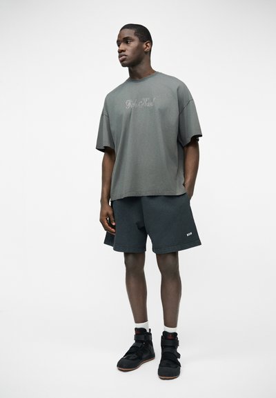 Jeune homme portant un T-shirt gris graphique oversize, un short ample foncé, des chaussettes blanches et des baskets montantes noires, debout les mains dans les poches.