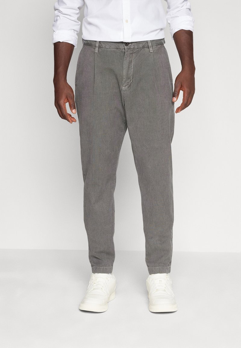 Homme portant un pantalon décontracté gris, une chemise blanche à manches retroussées et des baskets blanches, debout sur un fond blanc uni.
