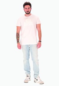 Uomo con barba e tatuaggi che indossa una t-shirt bianca Diesel, jeans azzurri chiari e sneakers bianche New Balance, in piedi contro uno sfondo bianco.