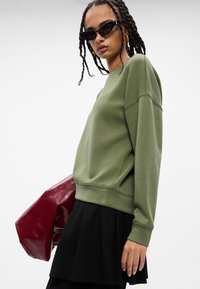 Sudadera verde con puños acanalados, combinada con una falda negra plisada. El modelo sostiene un bolso burdeos brillante y lleva unas gafas de sol negras elegantes.