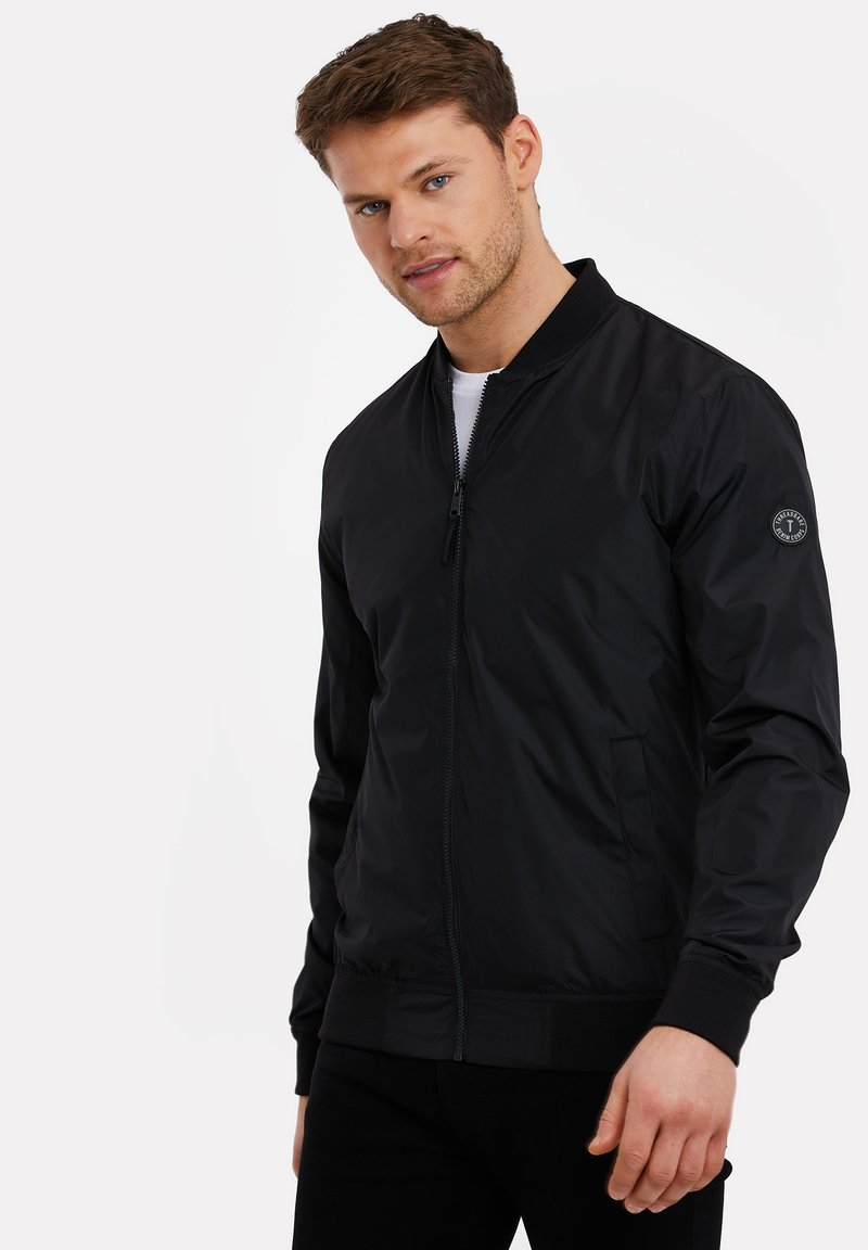 Schwarze Bomberjacke mit Reißverschluss an der Vorderseite, gerippten Bündchen und Saum. Glattes, leichtes Material mit Seitentaschen und einem Logo-Patch am Ärmel.