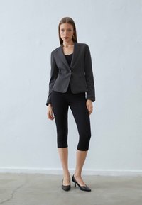 Nő áll egy sima falnak támaszkodva, szürke testhez simuló blézerben, fekete, rövid leggingsben és fekete magassarkú cipőben.