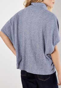 Hellblauer, gerippter Pullover mit hohem Kragen, kurzen Ärmeln und entspannter Passform, kombiniert mit dunkelblauen Jeans. Strukturiert gewebte Details sichtbar.