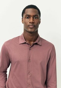 Jaunasis juodaodis vyras su trumpais garbanotais plaukais, vilkintis šviesiai rožinį marškinių su sagomis, stovi prieš šviesų vientisą foną.