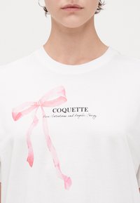 Vit T-shirt med en grafik av ett rosa band. Texten nedanför lyder "COQUETTE" och "Ren Avsikt och Änglalik Energi" i svart font.