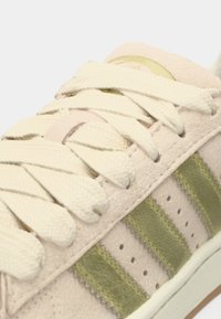 Μπεζ σουέτ αθλητικό παπούτσι με κρέμ κορδόνια, ρίγες σε ελιά, υφασμάτινες λεπτομέρειες και διάτρητη διακόσμηση στις πλευρές.