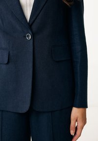 Navyblå skräddarsydd kavaj med strukturerade axlar, enknappsstängning och två framfickor, tillverkad av ett texturerat tyg.