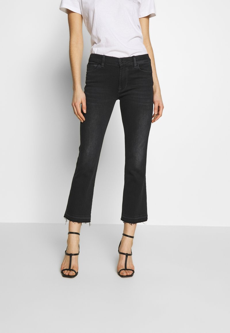 Femme portant un jean noir court avec des bords bruts, un t-shirt blanc, et des sandales à talons hauts noires à lanières, se tenant contre un fond blanc.