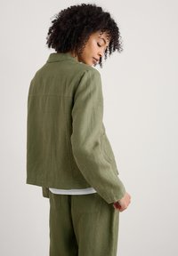 Chaqueta de lino ligera en verde oliva con cuello, bolsillos laterales y un corte relajado, vista desde la espalda con un conjunto a juego.
