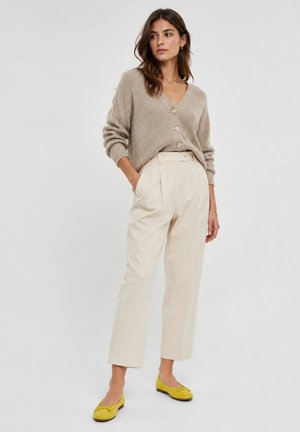 Woman wearing a beige buttoned cardigan, high-waist cream trousers with pleats, and yellow flats, posing with one hand in pocket.