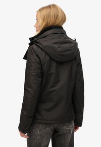 Veste matelassée noire avec capuche, fermeture éclair à l'avant et col côtelé. Présente une finition texturée avec un logo sur la manche. Portée avec un jean gris.