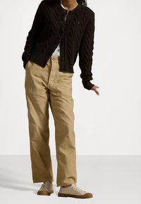 Brun kabelstickad cardigan, vit T-shirt, beige vida byxor och ljusbruna sneakers med strukturerad yta och bruna accenter.