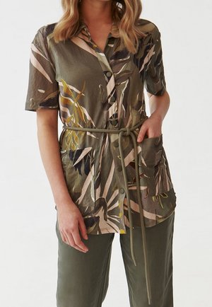 Woman wearing short-sleeve buttoned shirt with olive and beige floral print, belted waist, and large front pockets, paired with olive pants.