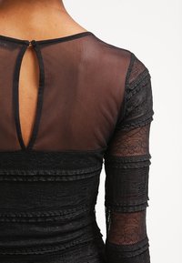 Robe noire avec un tissu texturé et superposé ainsi que des manches longues transparentes. Présente une ouverture en goutte d'eau au dos et des détails de couture délicats.