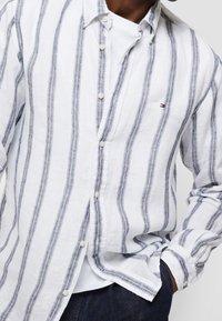 Camicia in lino bianco con righe verticali blu, chiusura frontale con bottoni e colletto standard. Presenta un piccolo logo sul petto e maniche lunghe.