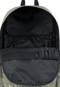 Interior de mochila de tela verde con forro negro. Las características incluyen un bolsillo con cremallera y compartimentos elásticos para la organización.