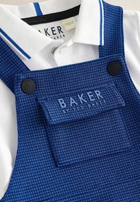 Blaue karierte Latzhose aus strukturiertem Stoff mit schwarzen Knöpfen und einer Tasche, die "BAKER BY TED BAKER" zeigt. Darunter ein weißes Polo.