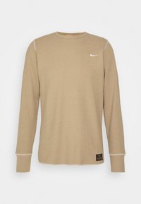 Chemise beige à manches longues avec une texture côtelée, un col rond et des surpiqûres blanches. Présente un petit logo Nike et une étiquette en tissu foncé à l'ourlet.