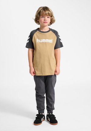 Ungt barn stående iført tan- og mørkegrå hummel-trøje, mørkegrå bukser og sorte sneakers med velcrolukning mod hvid baggrund.