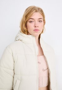 Giacca puffer beige chiaro con cappuccio, caratterizzata da una texture trapuntata e una zip frontale. Indossata sopra un top rosa testurizzato con logo.